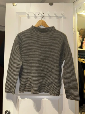 Grå ribbad tröja från H&M BASIC - Säljer en grå ribbad tröja från H&M BASIC i storlek S. Tröjan har ett diskret randigt mönster, högre krage och långa ärmar. Perfekt till lager på lager och enkel att matcha med olika stilar.