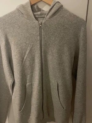 Grå core kofta från Jack & Jones - Grå hoodie från Jack & Jones Core i storlek S. Tröjan har huva, hel dragkedja framtill och två fickor. Mjuk och skön bomullsmix, perfekt för chill eller häng. Klassisk passform med långa ärmar och ribbade muddar. Hoodien är knappt använd och väldigt skön att använda under en höstlig dag. Kontakt för mer info! ABS! Pris går att diskutera!!!
