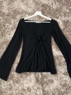 Svart topp från designbysi - Svart långärmad topp från Design by Si i storlek XS/S. Modellen har en smickrande V-ringning och en dekorativ knytdetalj framtill som ger fint fall och markerar midjan. Mjukt och stretchigt material som sitter bekvämt och följsamt på kroppen. Perfekt både till vardag och fest – lika fin till jeans som till kjol. Aldrig använd