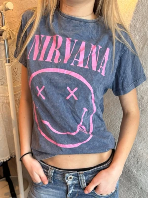 Blå Nirvana t-shirt med rosa tryck - Säljer en blå t-shirt med ikoniskt rosa Nirvana-tryck och smiley på bröstet. T-shirten har korta ärmar, klassisk passform och är gjord i mjuk bomull. 