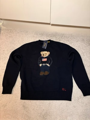 Mörkblå stickad tröja Polo Bear - Mörkblå stickad tröja från Polo Ralph Lauren med ikoniska Polo Bear-motivet på bröstet. Rund halsringning, ribbade muddar och RL-broderi i rött nedtill. Perfekt för dig som gillar klassisk preppy stil.