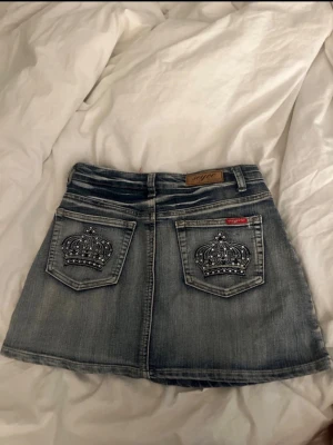Jeanskjol med kron-detaljer från Soya - Säljer en kort jeanskjol från Soya med snygga kronbroderier och nitar på bakfickorna. Kjolen har klassisk femficksmodell, knapp och dragkedja framtill samt en lätt utsvängd passform. Perfekt till sneakers eller boots. Nyskick