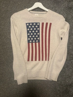 Beige stickad tröja med USA-flagga - Beige stickad tröja med stort tryck av amerikanska flaggan på bröstet. Tröjan har rund halsringning och långa ärmar. Perfekt för dig som vill sticka ut med en cool och klassisk look. Storlek 164