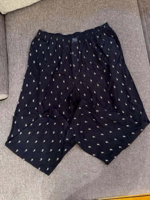 Mörkblå pyjamasbyxor från Polo Ralph Lauren storlek XL - Säljer ett par mörkblå pyjamasbyxor från Polo Ralph Lauren med klassiskt broderat polospelare-mönster i vitt över hela tyget. De har resår i midjan och knapp framtill. Perfekta för dig som gillar stilrena och bekväma kläder