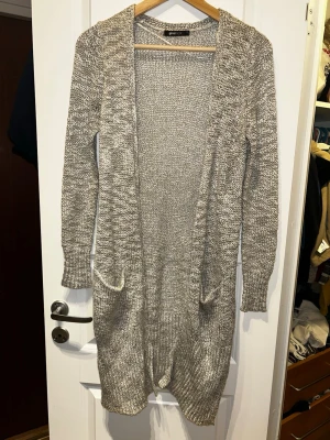 Lång stickad kofta från Gina Tricot - Lång, mysig stickad kofta i melerad grå färg från Gina Tricot. Koftan har två fickor framtill, ribbade muddar och slitsar nedtill. Perfekt att slänga på sig över en t-shirt eller topp när det är kyligt ute.