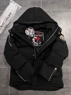 Svart pufferjacka från Moose Knuckles - Säljer en svart pufferjacka från Moose Knuckles med huva och feta dragkedjor. Jackan har en broderad etikett på insidan och metallogo på ärmen. Perfekt för kalla dagar och riktigt snygg streetstil.