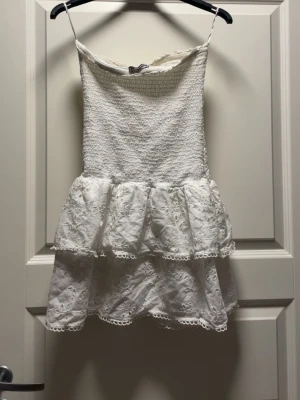 Vit kort klänning med volanger från Gina tricot - Supersöt vit kort klänning med smockad överdel och dubbla volanger i kjolen. Klänningen har tunna axelband och broderade spetsdetaljer på kjolen. Perfekt för sommarens alla tillfällen och ger en romantisk vibe.