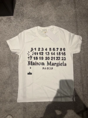 Vit Maison Margiela t-shirt med print - Vit t-shirt från Maison Margiela med svart pixelinspirerat tryck och logga framtill. Klassisk rund hals och korta ärmar. Tillverkad i mjuk bomull för en skön känsla. Perfekt för dig som gillar unika designer och streetwear.