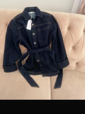 Mörkblå jeansjacka med bälte Sanne Alexandra - Snygg mörkblå jeansjacka från Sanne Alexandra med knappar framtill, bröstficka och tillhörande bälte i midjan. Jackan har klassisk krage, långa ärmar och kontrastsömmar som ger en cool look. Perfekt att styla med jeans eller kjol för en trendig outfit. Org pris: 1195 kr