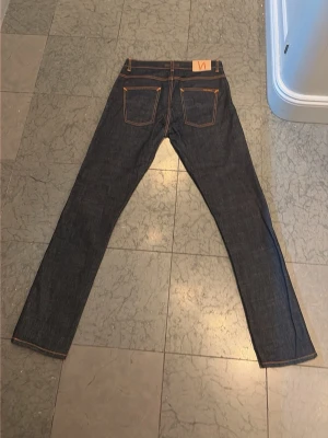 Nudie Jeans blå jeansbyxor W32 L34 - Säljer ett par mörkblå jeans från Nudie Jeans i klassisk rak modell. Byxorna har kontrastsömmar i orange, fem fickor och Nudie-logga på bakfickan. Tillverkade i slitstarkt jeansmaterial, perfekta för dig som gillar stilrena och tidlösa jeans.