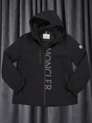 Moncler vind jacka  - Säljer en svart vindjacka från Moncler med huva och dragkedja framtill. Jackan har stora Moncler-trycket längs bröstet och klassisk logga på ärmen. Två fickor med dragkedja och elastiska muddar vid ärmsluten. Perfekt för dig som vill ha en stilren och sportig look.