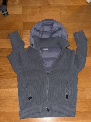 Grå moncler stickad cardigan  - Tjena! Säljer en grå stickad cardigan från Moncler med stickade ärmar och huva. Jackan har dragkedja framtill, två fickor med dragkedja och Moncler-logga på ena ärmen. Baksidan och huvan är quiltad och vadderad för extra värme. Den har en liten defekt som kan visas vid intresse hålet är ca 2 milmeter och jätte enkelt att laga . Perfekt för kyliga dagar, samt för normala dagar. Pris kan diskuteras vid snabb och smidig affär! Hör av dig vid frågor 