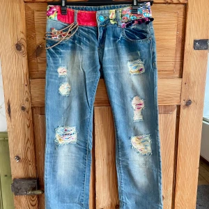 Desigual jeans Stl 36 - Unika blå jeans från Desigual med färgglada blommiga tygpatchar, träpärlor och broderade detaljer. Slitna partier med lappar i olika färger och mönster, samt midja dekorerad med rosa och grönt tyg. Rak passform och coola detaljer som gör dem till ett statement-plagg.
