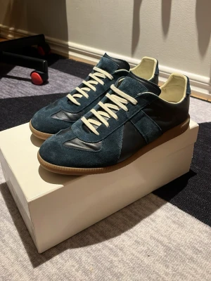 Maison Margiela (äkta) - Pris kan diskuteras, Säljer mina  Maison Margiela gats sneakers i mörkblå mocka och skinn jävligt feta, köpta i butik med vita snören och beige innersida. Skorna har klassisk gummisula och stilren design, perfekta för dig som gillar exklusiva och snygga sneakers.