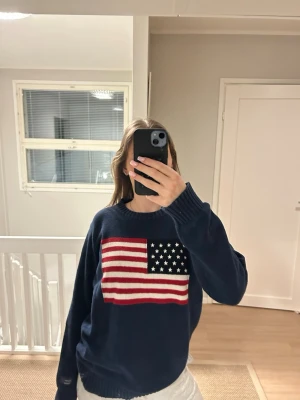 Mörkblå stickad tröja med USA-flagga - Säljer en mörkblå stickad tröja med stor amerikansk flagga på bröstet. Tröjan har rund halsringning, ribbade muddar och lång ärm. Perfekt för dig som vill ha en statement piece i garderoben!