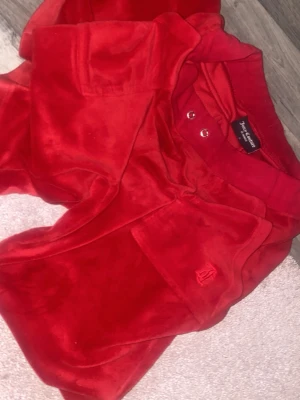 Röda mjukisbyxor Juicy Couture - Säljer ett par ikoniska röda mjukisbyxor från Juicy Couture i mjuk sammet. Byxorna har normal passform, raka ben och resår i midjan med metallöljetter. Klassisk Juicy-brodyr på fickan framtill. Perfekta för en chill och trendig look. Band saknas 