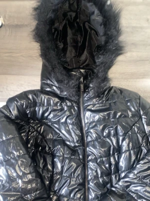 Svart glansig pufferjacka med fuskpäls - Säljer en svart, glansig pufferjacka med huva och fuskpäls runt kanten. Jackan har quiltad design och dragkedja framtill. Perfekt för kalla dagar när du vill hålla dig varm och ändå se snygg ut.