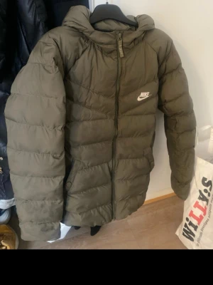 Olivgrön pufferjacka från Nike M - Säljer en olivgrön pufferjacka från Nike i storlek M.Jackan har huva, dragkedja framtill och klassisk Nike-logga på bröstet. Tillverkad i syntetmaterial och perfekt för kalla dagar. Passar dig som gillar en sportig och clean stil.