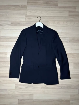 Mörkblå kostym från Riley - Säljer en stilren mörkblå kostym från Riley. Klassisk modell med två fickor fram, bröstficka och knappar vid ärmslut. Fodrad insida i glansigt tyg och smal passform. Perfekt för dig som vill ha en clean och tidlös look.