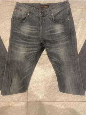 Gråa Nudie Jeans - Säljer ett par gråa jeans från Nudie Jeans i storlek W27 L32. Jeansen har klassisk femficksdesign, svarta sömmar och subtila slitningar framtill för en cool look. Tillverkade i mjukt jeansmaterial med stretch för extra komfort.