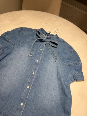 Blå jeansskjorta med knyt från Zara - Säljer en blå jeansskjorta från Zara i storlek M. Skjortan har korta ärmar, tryckknappar framtill och en snygg knyt-detalj i halsen. Tillverkad i bomull för en chill och avslappnad look.