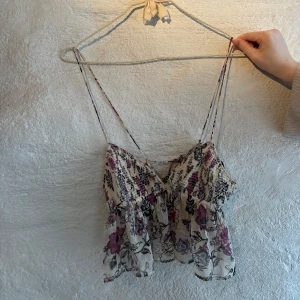 Blommig linnetopp med volang - Supersöt linnetopp med blommigt mönster i lila, svart och beige. Toppen har tunna axelband som går att ljustera, volang nertill och smockad överdel. Perfekt för varma dagar och ger en romantisk vibe.