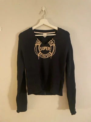 Svart stickad tröja Marc O'Polo S - Svart stickad tröja från Marc O'Polo Campus i storlek S. Tröjan har v-ringning och beige broderad text 'SUPER FINISH' framtill. Ribbstickade muddar vid ärmslut och nederkant ger en snygg passform. Perfekt till höst och vinter!
