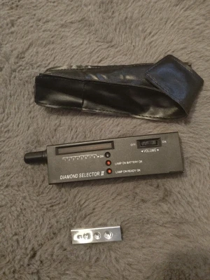 Diamond tester - Bilden visar en Diamond Selector II, ett verktyg för att testa äktheten på diamanter, samt ett svart fodral och ett metallstycke. 