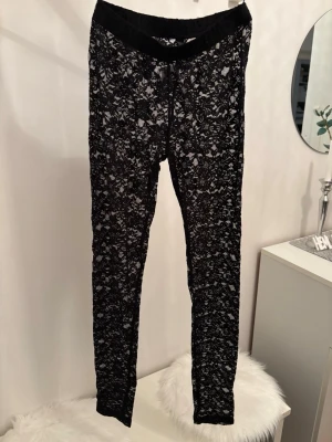 Svarta spetsleggings med blommönster - Säljer ett par svarta leggings i stretchigt material med snyggt blommigt spetsmönster. De har en bred resår i midjan för extra komfort och sitter tight längs benen. Perfekta för dig som vill ha något unikt och trendigt i garderoben.