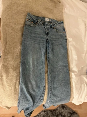 Broderade ljusblå jeans från 157 - Säljer ett par ljusblå jeans från 157 med snygg broderad detalj i guld på bakfickorna. Klassisk femficksmodell med raka ben och dubbla knappar i midjan. Jeansen har en relaxed passform och är tillverkade i mjukt bomullsjeans-tyg.