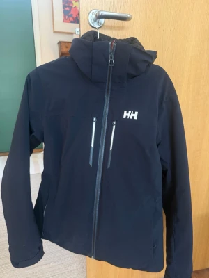 Svart vindjacka från Helly Hansen L - Säljer en svart vindjacka från Helly Hansen i storlek L. Jackan har huva, två bröstfickor med dragkedja och hel dragkedja framtill. Materialet är slitstarkt och vattenavvisande syntet, perfekt för blåsiga dagar. Klassisk HH-logga på bröstet och quiltat innerfoder.