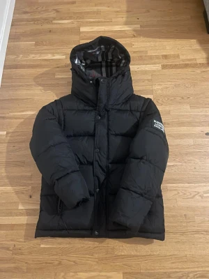 Svart pufferjacka från Burberry - Säljer en svart pufferjacka från Burberry med klassiskt rutigt foder i huvan och tryckt logga på ärmen. Jackan har dragkedja och knappar framtill, två fickor med dragkedja och en hög krage. Perfekt för kalla dagar och riktigt snygg streetstil.