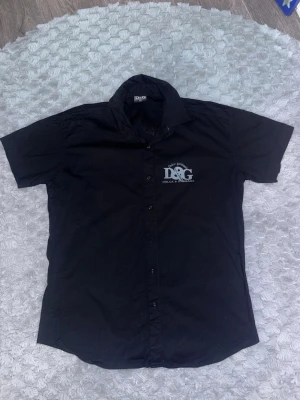 Svart skjorta Dolce & Gabbana - Svart skjorta från Dolce & Gabbana i storlek XXL. Skjortan har stora vita D&G-loggor tryckta på bröstet och ryggen. Klassisk och knäppare. Tillverkad i bomull för en skön känsla och stilren look. skriv gärna för mer frågor och bilder på skjortan så löser jah