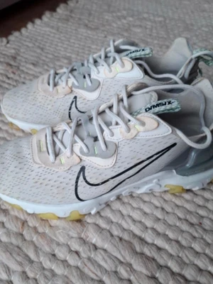 Nike React sneakers i ljusgrått - Nike React sneakers i ljusgrå mesh med vita och beige detaljer samt svart swoosh på sidan. Skorna har snörning, platt sula med gula inslag och en modern, sportig design. Perfekta för dig som gillar streetstyle och bekväma skor med cool look.