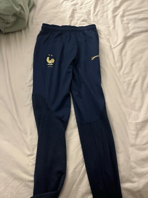 Marinblå Nike FFF träningsbyxor storlek L i barn alltså 147-158 - Säljer ett par marinblå träningsbyxor från Nike med FFF-logga och två guldstjärnor. Byxorna har guldfärgad Nike Swoosh, smal passform och är tillverkade i Dri-FIT material som andas. Perfekta för fotbollsträning eller chill.      ….. pris kan diskuteras!