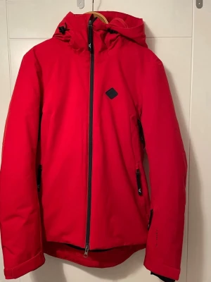 Röd vindjacka från J.Lindeberg - Starling skidjacka från J Lindeberg för skidåkning och vinteraktiviteter med högt tempo.  Jackan kombinerar teknisk funktion med stilren passform. Med 20K-vattentäthet, vindtätt yttertyg och Primaloft® Silver-isolering håller du dig varm och torr i tuffa förhållanden. Den slimmade passformen ger ett följsamt intryck – välj en storlek större om du vill ha mer rörelseutrymme. Ventilation under armarna och snöskydd i midjan ger ökad komfort i varierat väder, medan den dolda liftkortsfickan och just