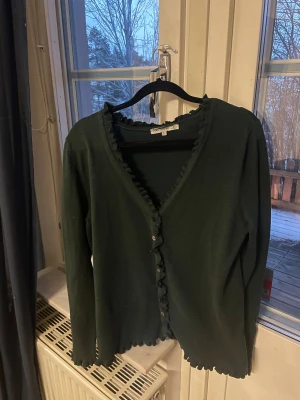 Mörkgrön kofta med volang från Flash Woman - Snygg mörkgrön kofta från Flash Woman i storlek L. Koftan har volangdetaljer längs v-ringningen och nederkanten, samt knappar framtill. Långärmad och tillverkad i mjukt stickat material som känns skönt mot huden. Det står storlek L men den passar mer en M.