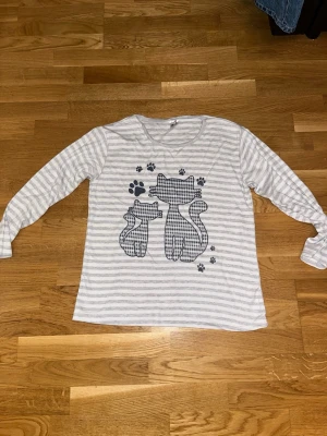 Randig långärmad tröja Miss Lale - Långärmad tröja från Miss Lale i vitt och grått med tunna ränder. Framsidan har ett gulligt kattmotiv och tassavtryck i mörkblått. Tröjan har rund halsringning och är gjord i mjukt bomullsmaterial. Perfekt för dig som gillar söta detaljer!