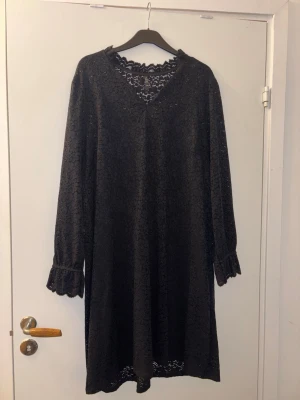 Svart spetsklänning från LUX - Svart klänning från LUX i storlek 2XL, helt täckt av blommig spets. Klänningen har v-ringning, långa ärmar med volangkant och fina spetsdetaljer vid hals och ärmslut. Perfekt för dig som gillar en elegant och feminin stil.