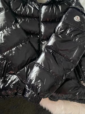 Svart pufferjacka från Moncler - Säljer en svart, glansig pufferjacka från Moncler med klassisk logga på ärmen. Jackan har dragkedja framtill, flera fickor med dragkedja och quiltad design. Perfekt för kalla dagar och riktigt snygg till streetwear-stil.