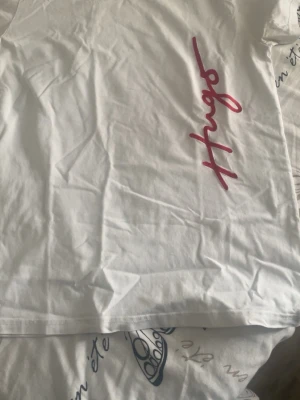 Vit t-shirt från Hugo med röd logga - Säljer en clean vit t-shirt från Hugo med röd logga tryckt vertikalt på framsidan. Klassisk rund hals och korta ärmar. Skön och mjuk i bomull, perfekt till jeans eller shorts. Enkel men ändå snygg detalj med loggan.