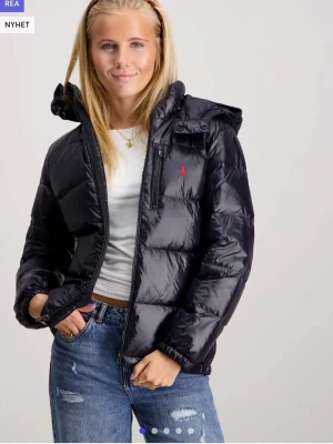 Svart pufferjacka från Ralph Lauren - Snygg svart pufferjacka från Ralph Lauren med röd logga på bröstet. Jackan har huva, dragkedja framtill och en bröstficka med dragkedja. Perfekt för kyliga dagar och riktigt skön att bära över en hoodie eller t-shirt. Storlek XL BARN vilket ungefär motsvarar storlek S, från kidsbrandstore.