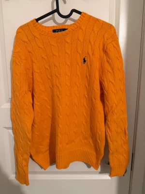  tröja Polo Ralph Lauren - Säljer en orange kabelstickad tröja från Polo Ralph Lauren i storlek M. Tröjan har rund halsringning, ribbade muddar och den klassiska blå broderade loggan på bröstet. Perfekt för dig som vill ha en färgklick i garderoben.