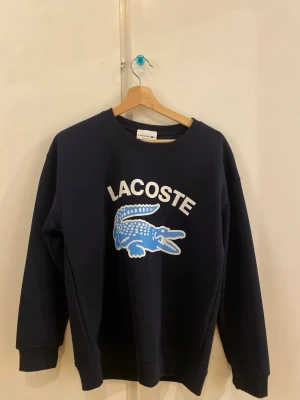 Mörkblå sweatshirt från Lacoste - Snygg mörkblå sweatshirt från Lacoste med stor vit logga och blå krokodil framtill. Tröjan har rund hals, långa ärmar och regular fit. Perfekt för dig som gillar klassisk streetstyle och vill ha något bekvämt och stilrent.