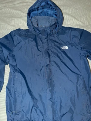 Blå vindjacka från The North Face - Säljer en blå vindjacka från The North Face med huva och dragkedja framtill. Jackan har två sidofickor och meshfoder på insidan för extra komfort. Perfekt för blåsiga dagar och enkel att packa ner i väskan.