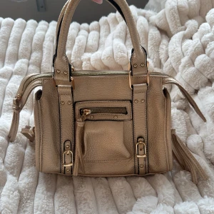 Beige handväska med dragkedja och detaljer - Snygg beige handväska i skinnimitation med både handtag och axelrem. Väskan har coola spänndetaljer, dragkedja framtill och dekorativa tofsar. Perfekt storlek för att få plats med allt du behöver och passar till många olika outfits.