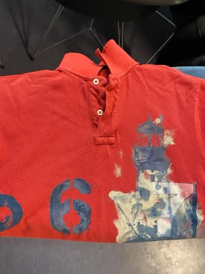 Röd henley från Polo Ralph Lauren - Röd henley-skjorta från Polo Ralph Lauren i bomull. Framsidan har ett stort tryck med siffror, fyr och text i blått och vitt. Skjortan har korta ärmar, två knappar vid halsen och klassisk krage.