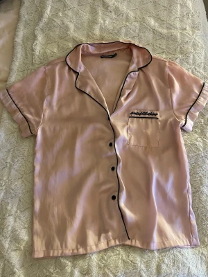 Rosa satinpyjamas från PrettyLittleThing - Säljer ett set med kortärmad pyjamasskjorta och shorts i ljusrosa satin från PrettyLittleThing. Nån lös tråd