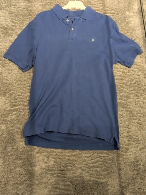 Blå piké med liten logga - Säljer en klassisk blå pikétröja med krage och två knappar framtill. Tröjan har korta ärmar och en liten broderad logga på bröstet i grönt och gult. Perfekt till jeans eller shorts för en chill look.