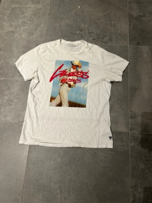 Vit t-shirt med tryck från Guess - Vit t-shirt från Guess med stort färgglatt tryck framtill och röd text. Tillverkad i 100% ekologisk bomull och har klassisk passform med korta ärmar. Perfekt för dig som gillar streetstyle och vill sticka ut lite extra.                                            Info: det finns något pyttelitet hål på tröjan men som inte märks. Annars är den super!